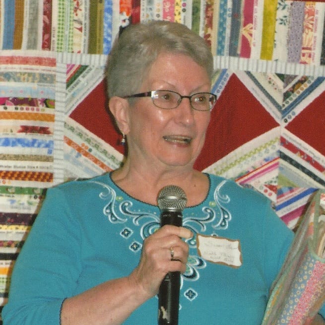 Susan Goins, quiltmaker and seamstress, holding a microphone, standing in front of a colorful quilt